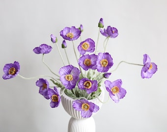 May include: A bouquet of purple silk poppy flowers in a white vase. The flowers have yellow centers and are arranged in a vase with a textured surface.