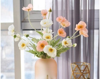 May include: A bouquet of delicate pink and white poppy flowers arranged in a frosted pink glass vase. The vase is sitting on a white table with a gold decorative object in the background.