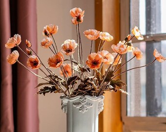 May include: A white ceramic vase with a decorative rim holds a bouquet of orange poppy flowers. The vase sits on a wooden table next to an open book, a small bowl, and a mug.