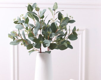Può includere: Un vaso in ceramica bianca con una texture a coste contiene un bouquet di rami di eucalipto artificiali con foglie verdi.