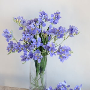 Puede incluir: Un ramo de flores artificiales de fresia de color lavanda en un jarr&oacute;n de vidrio transparente. Las flores est&aacute;n dispuestas en un estilo suelto y natural.