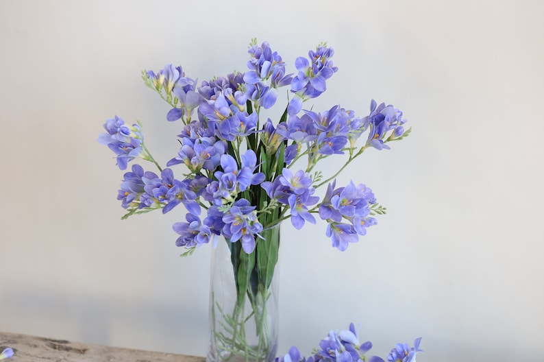 Puede incluir: Un ramo de flores artificiales de fresia de color lavanda en un jarr&oacute;n de vidrio transparente. Las flores est&aacute;n dispuestas en un estilo suelto y natural.