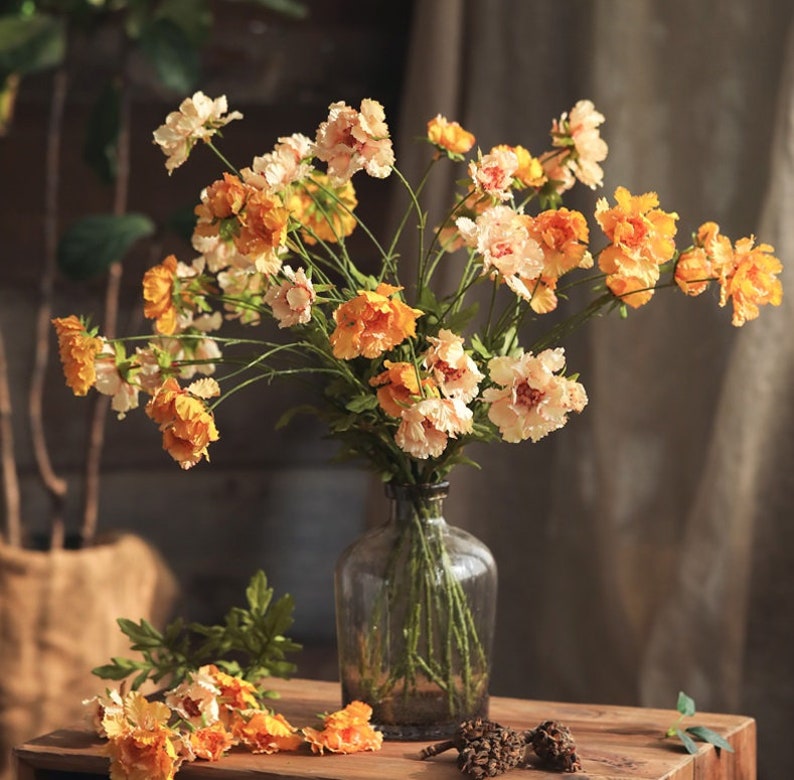May include: A clear glass bottle vase filled with a bouquet of artificial flowers in shades of orange, yellow, and cream. The flowers feature ruffled petals and thin green stems. Several loose flower heads and two small pinecones rest on the surface.