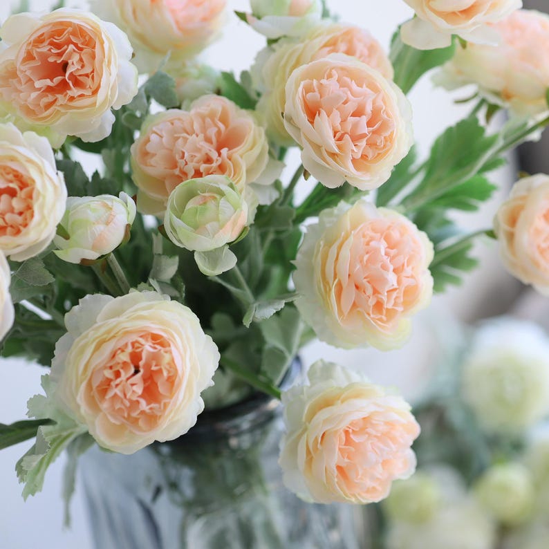 Puede incluir: Un ramo de flores artificiales de color melocot&oacute;n con una textura suave y rizada. Las flores est&aacute;n dispuestas en un jarr&oacute;n de vidrio transparente con una superficie texturizada.