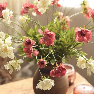 May include: A bouquet of artificial flowers in a brown vase, featuring a mix of ruffled white and deep pink blossoms on thin green stems with small green leaves. The flowers are arranged in a rounded cluster, displaying varied petal textures.