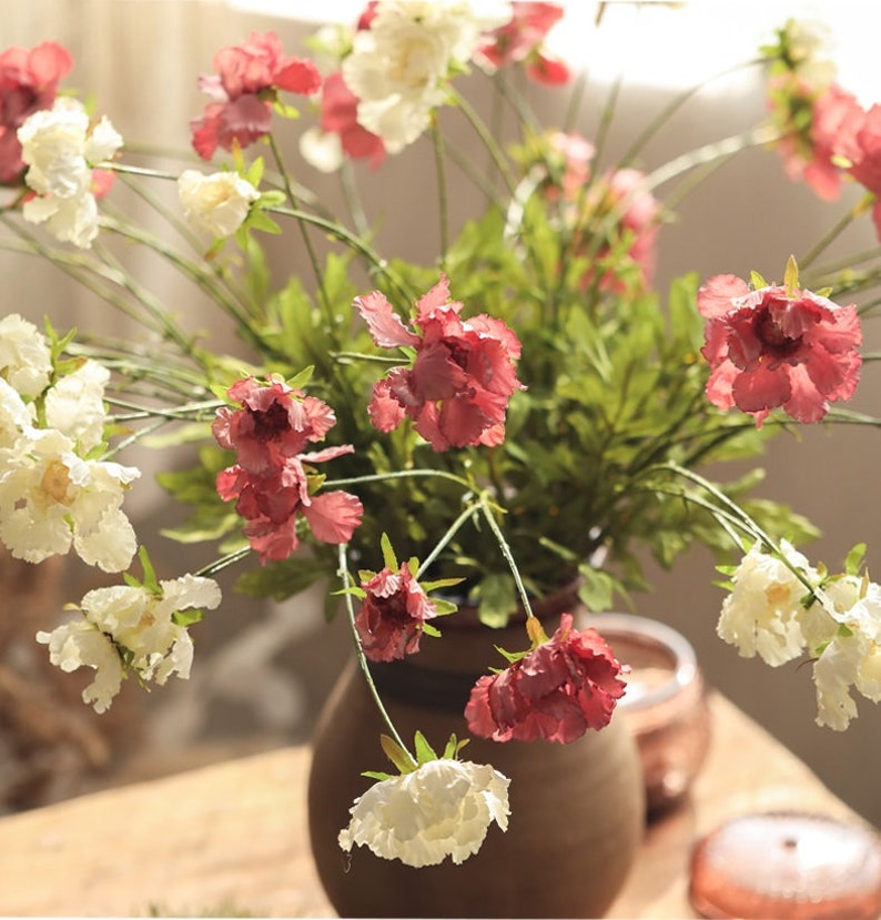 May include: A bouquet of artificial flowers in a brown vase, featuring a mix of ruffled white and deep pink blossoms on thin green stems with small green leaves. The flowers are arranged in a rounded cluster, displaying varied petal textures.