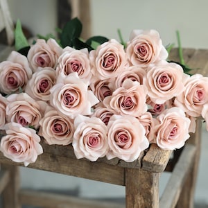 Puede incluir: Un ramo de rosas artificiales de color rosa claro dispuestas sobre una silla de madera. Las rosas est&aacute;n hechas de un material suave y sedoso y tienen un aspecto realista.