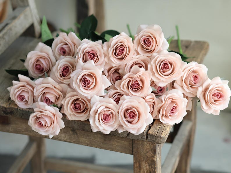 Puede incluir: Un ramo de rosas artificiales de color rosa claro dispuestas sobre una silla de madera. Las rosas est&aacute;n hechas de un material suave y sedoso y tienen un aspecto realista.