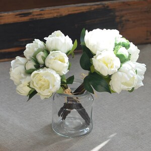 May include: A bouquet of white peonies with green leaves in a clear glass vase.
