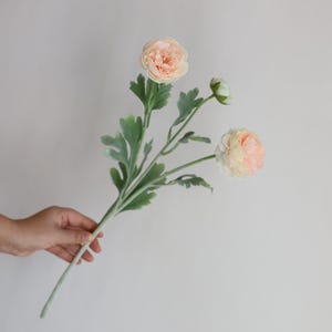 Puede incluir: Un ramo de tres flores de ran&uacute;nculo artificiales de color melocot&oacute;n con hojas verdes. Las flores est&aacute;n dispuestas en un ramo sostenido en la mano.