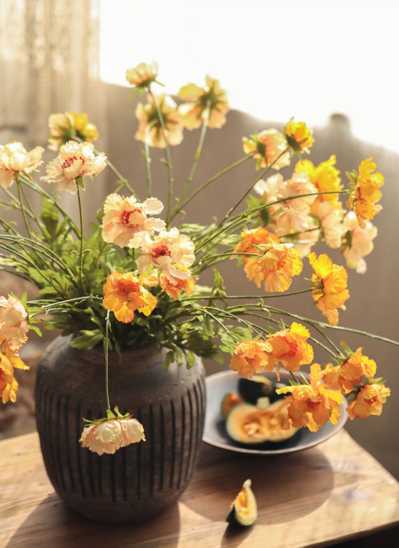 May include: A dark brown, vertically ribbed ceramic vase filled with a bouquet of yellow and orange ruffled flowers on thin green stems. Beside the vase sits a shallow bowl containing sliced green squash and a single piece of squash.