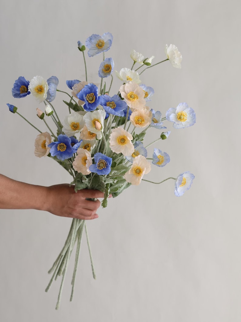 Puede incluir: Un ramo de flores de amapola de seda azul y crema con centros amarillos. Los tallos son verdes y tienen una textura peluda.
