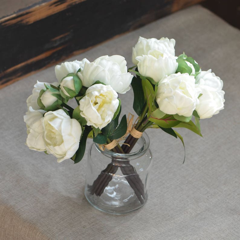 May include: A bouquet of white peonies arranged in a clear glass vase. The peonies have green leaves and stems.
