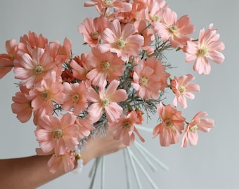 May include: A bouquet of peach-colored cosmos flowers with yellow centers and green accents. The flowers are held together by white stems, creating a delicate and elegant floral arrangement. Perfect for home decor or special events.