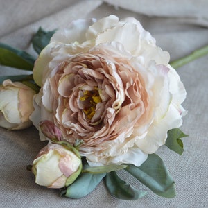 May include: A bouquet of artificial peonies featuring a single large, blush-coloured peony and two smaller, cream-coloured peonies. The peonies are arranged with green leaves on a light brown linen background.