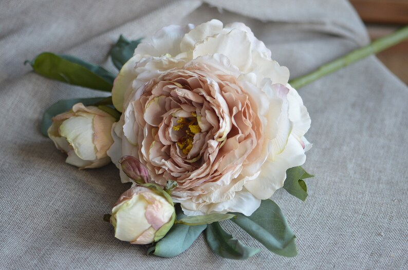 May include: A bouquet of artificial peonies featuring a single large, blush-coloured peony and two smaller, cream-coloured peonies. The peonies are arranged with green leaves on a light brown linen background.