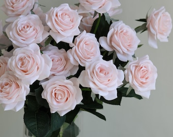 May include: A bouquet of light pink roses in a clear glass vase. The roses have multiple layers of delicate petals and green leaves. The vase is filled with water and the stems of the roses are visible.