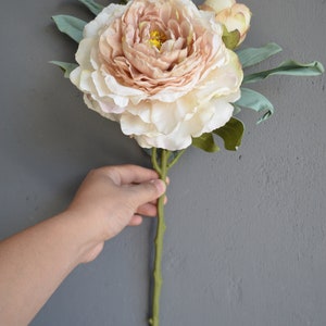 May include: A single artificial peony flower in a light pink and cream colour. The flower has a green stem and leaves.