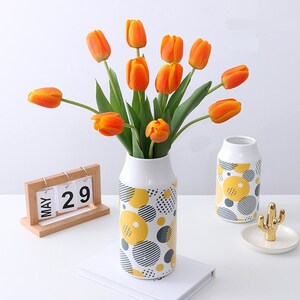 May include: A bouquet of orange tulips in a white ceramic vase with a yellow and black geometric pattern. The vase is sitting on a white surface with a wooden calendar that says "MAY 29".