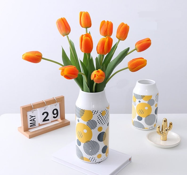 May include: A bouquet of orange tulips in a white ceramic vase with a yellow and black geometric pattern. The vase is sitting on a white surface with a wooden calendar that says "MAY 29".