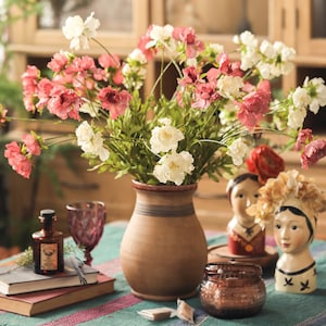 May include: A brown ceramic vase filled with white and pink flowers, positioned next to a small amber glass bottle, two stacked books, a purple glass, a lidded amber glass jar, and two small ceramic head sculptures on a green striped fabric.