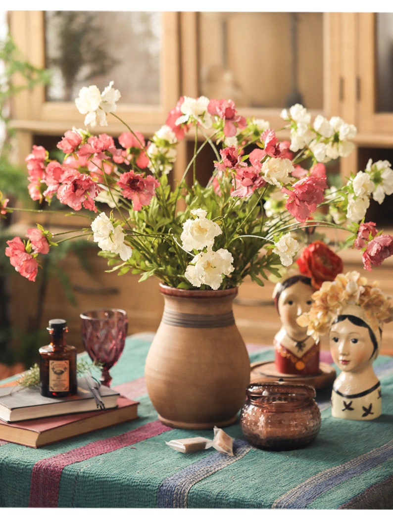 May include: A brown ceramic vase filled with white and pink flowers, positioned next to a small amber glass bottle, two stacked books, a purple glass, a lidded amber glass jar, and two small ceramic head sculptures on a green striped fabric.