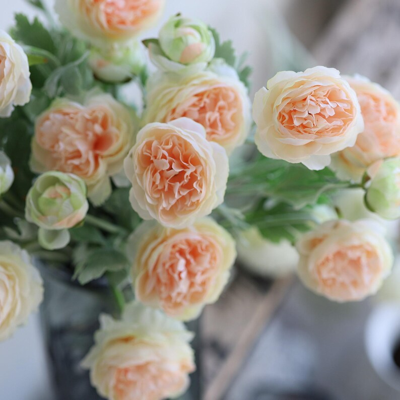 Puede incluir: Un ramo de flores artificiales de color melocot&oacute;n con hojas verdes. Las flores est&aacute;n dispuestas en un jarr&oacute;n.