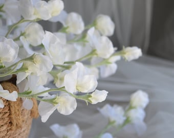 May include: Close-up of white sweet pea flowers in a woven basket. The delicate petals are a soft white, with green stems. The flowers are arranged in a natural, organic style, perfect for home decor or special events.