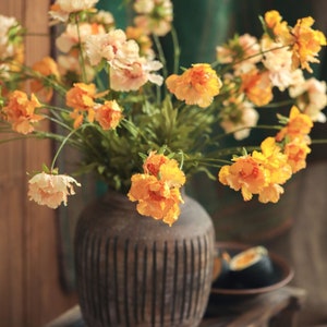 May include: A bouquet of artificial flowers in shades of orange, yellow, and cream, featuring ruffled petals and green stems, arranged in a dark brown, round, vertically ribbed ceramic vase with a textured, matte finish.