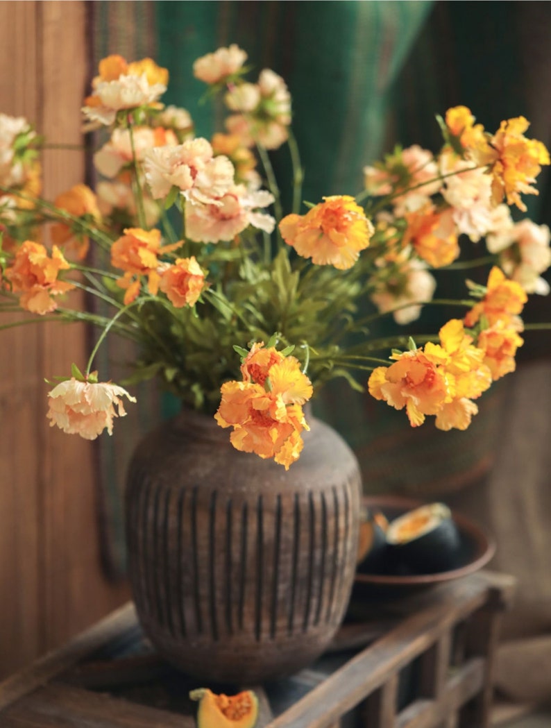 May include: A bouquet of artificial flowers in shades of orange, yellow, and cream, featuring ruffled petals and green stems, arranged in a dark brown, round, vertically ribbed ceramic vase with a textured, matte finish.