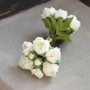 May include: Two bouquets of white artificial peonies with green leaves. The peonies are arranged in a cluster and have a realistic appearance.