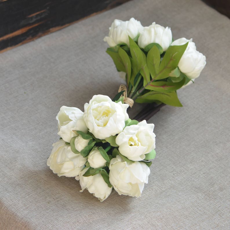 May include: Two bouquets of white artificial peonies with green leaves. The peonies are arranged in a cluster and have a realistic appearance.