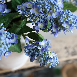 May include: A bouquet of artificial blue hydrangea flowers with green leaves. The flowers are arranged in a white vase.