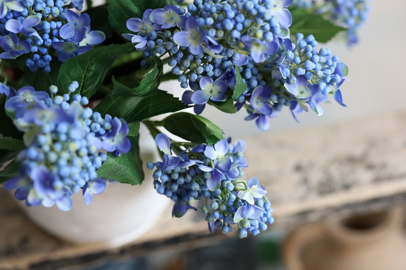 May include: A bouquet of artificial blue hydrangea flowers with green leaves. The flowers are arranged in a white vase.