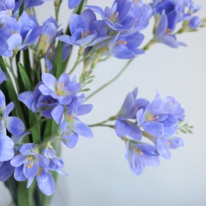 Puede incluir: Un ramo de flores de fresia de color lila claro en un jarr&oacute;n de vidrio transparente. Las flores est&aacute;n dispuestas en un estilo suelto y natural.