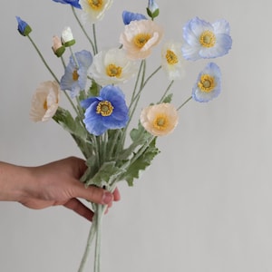 Puede incluir: Un ramo de flores de amapola artificiales en tonos azules, blancos y melocot&oacute;n. Las flores tienen centros amarillos y est&aacute;n dispuestas en un ramo sostenido en la mano.