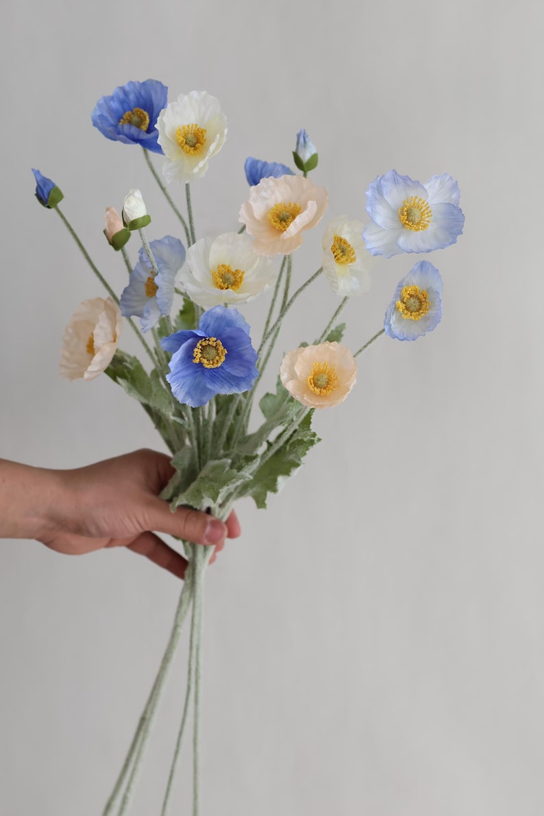 Puede incluir: Un ramo de flores de amapola artificiales en tonos azules, blancos y melocot&oacute;n. Las flores tienen centros amarillos y est&aacute;n dispuestas en un ramo sostenido en la mano.