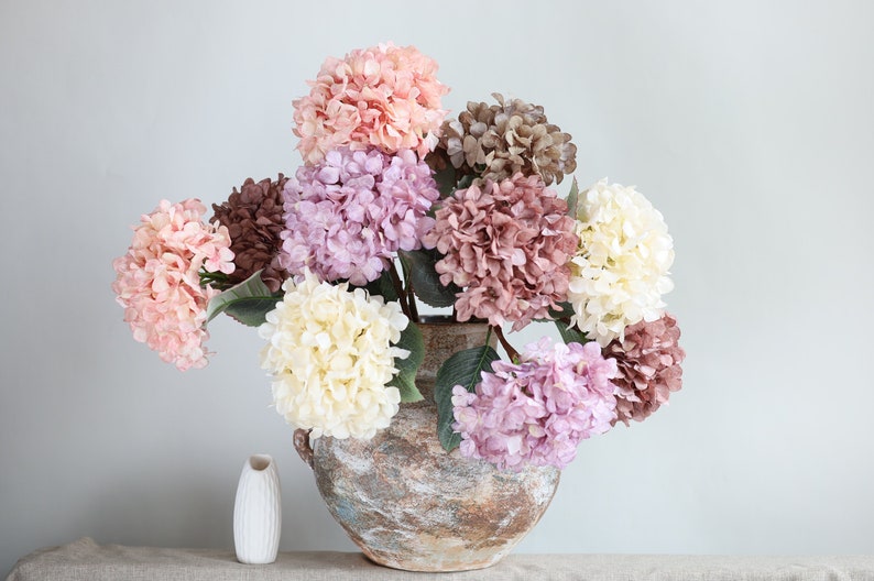 Puede incluir: Un ramo de hortensias artificiales en un jarr&oacute;n marr&oacute;n r&uacute;stico. Las flores son en tonos de rosa, morado y blanco.