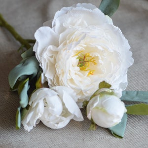 May include: A bouquet of three white peonies with green leaves. The peonies are in various stages of bloom, with one fully open and two in bud.