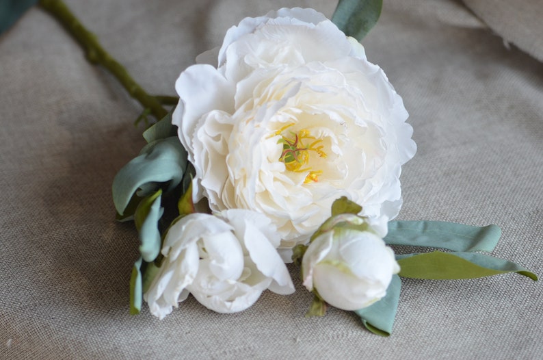 May include: A bouquet of three white peonies with green leaves. The peonies are in various stages of bloom, with one fully open and two in bud.