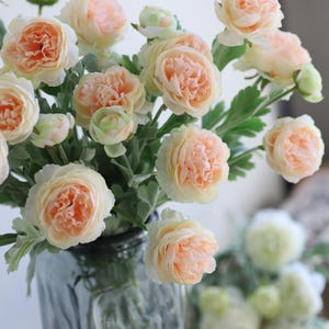Puede incluir: Un ramo de flores artificiales de color melocot&oacute;n con hojas verdes en un jarr&oacute;n de vidrio transparente. Las flores est&aacute;n dispuestas en un racimo y tienen un aspecto delicado y arrugado.