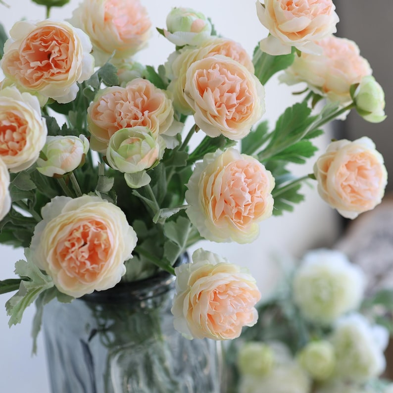 Puede incluir: Un ramo de flores artificiales de color melocot&oacute;n con hojas verdes en un jarr&oacute;n de vidrio transparente. Las flores est&aacute;n dispuestas en un racimo y tienen un aspecto delicado y arrugado.