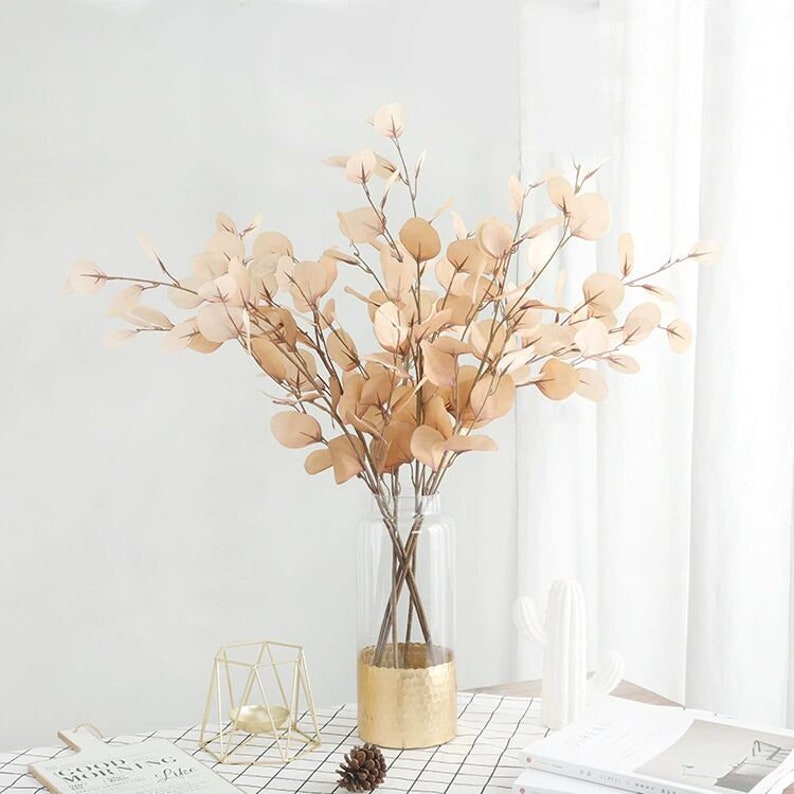 May include: A bouquet of dried eucalyptus branches in a clear glass vase with a gold rim. The vase is sitting on a white and black checkered table cloth.