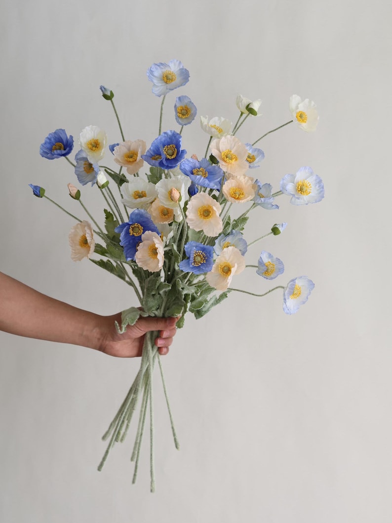 Puede incluir: Un ramo de flores de amapola de seda azul, blanco y melocot&oacute;n con tallos verdes.