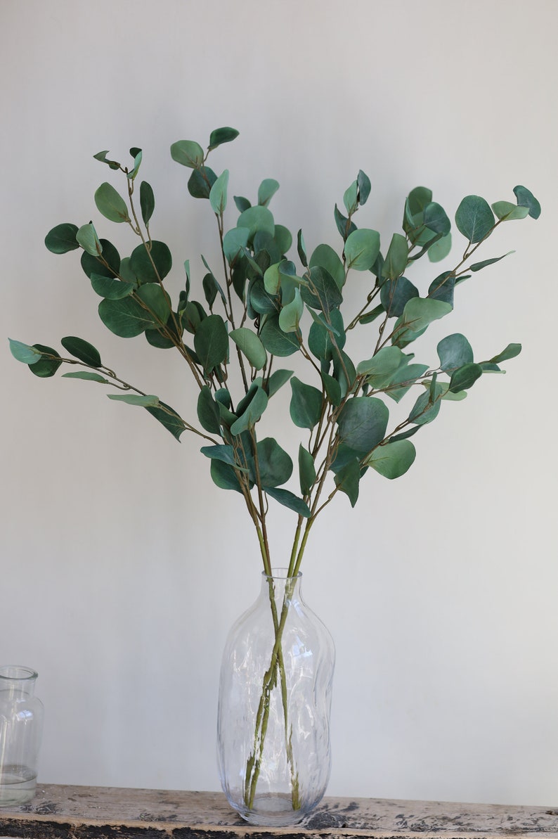 Op de afbeelding: Een bos kunstmatige eucalyptus takken in een heldere glazen vaas met een textuur. De vaas staat op een houten oppervlak.