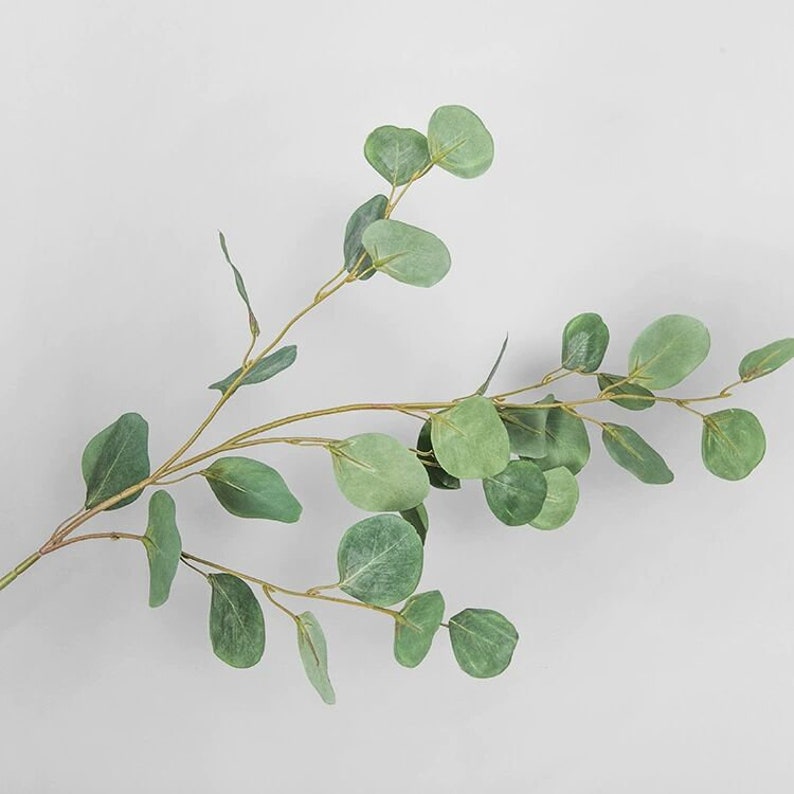 May include: A single stem of artificial eucalyptus with green leaves. The leaves are oval shaped and have a slightly textured surface.