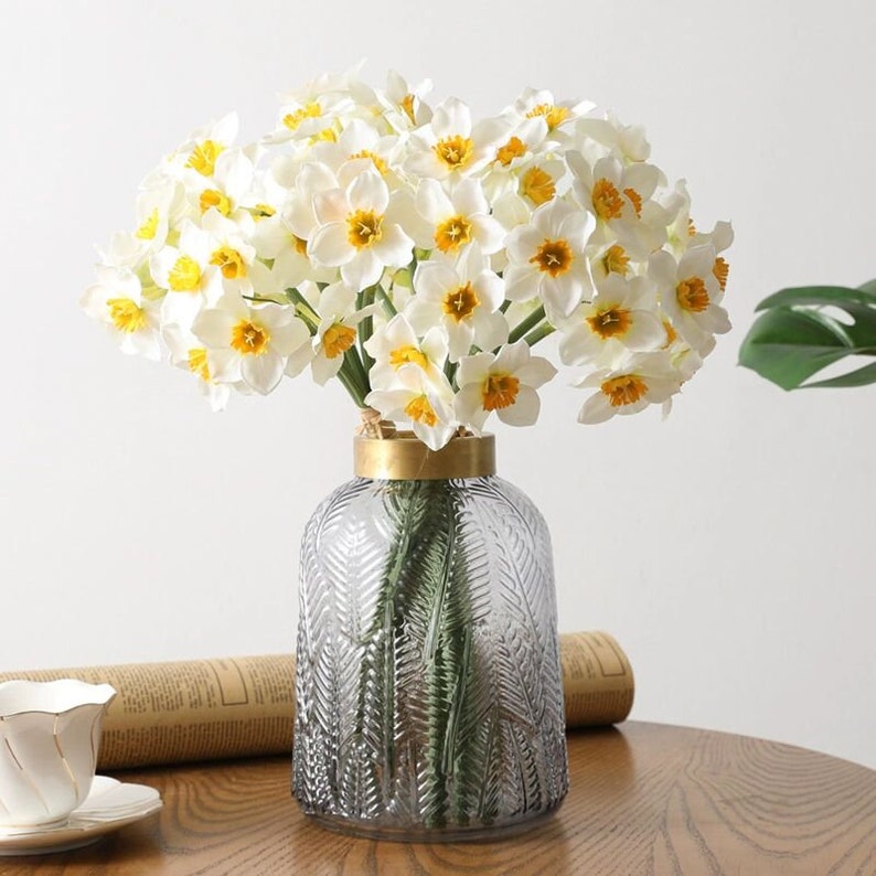 Peut inclure: Un bouquet de jonquilles blanches avec des centres jaunes dispos&eacute; dans un vase en verre gris avec un bord dor&eacute;. Le vase a un motif textur&eacute; de feuilles.