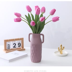 May include: A bouquet of pink tulips in a textured purple vase with two handles. The vase is on a white surface with a wooden calendar that says "MAY 29" and a small gold cactus on a white dish.
