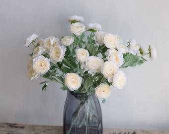 May include: A bouquet of cream-colored artificial ranunculus flowers in a gray vase. The flowers have a delicate, layered appearance and are arranged in a loose, natural style.