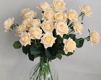 May include: A bouquet of cream-colored artificial roses arranged in a clear glass vase. The roses are clustered together and have a realistic appearance.